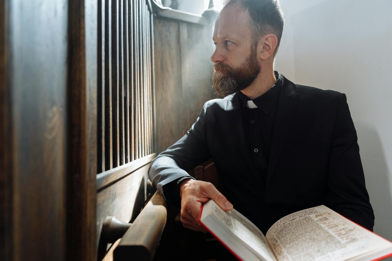 A bearded priest sits in a church confessional, reading a Bible, capturing a moment of quiet reflection and faith.